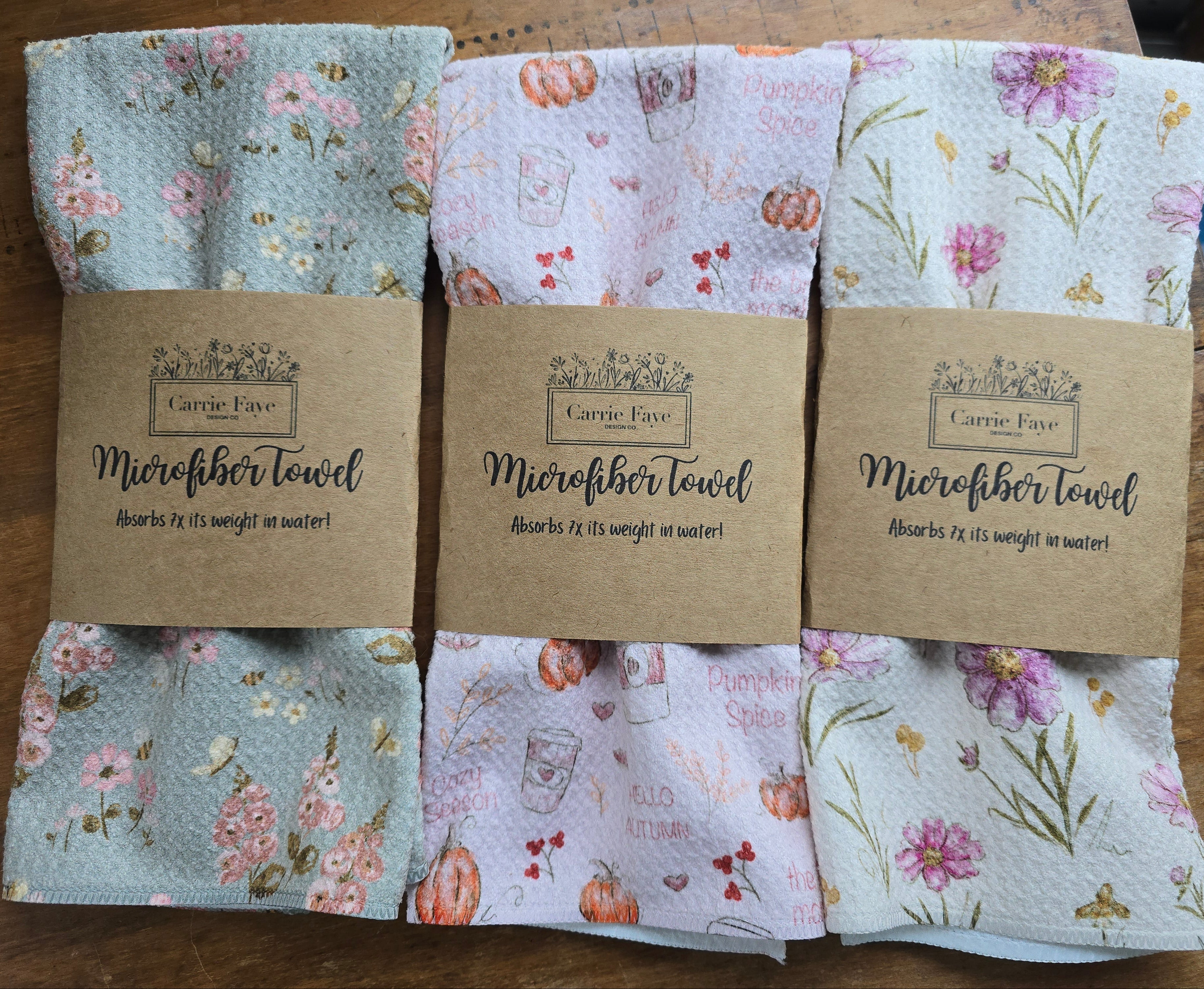 Three folded microfiber towels with floral and pumpkin patterns, each wrapped in a brown paper band labeled Carrie Fay Microfiber Towel, displayed on a wooden surface.
