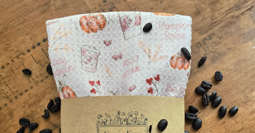 A folded microfiber towel with a fall-themed print of pumpkins, coffee cups, and text like 'Pumpkin Spice' and 'Hello Autumn,' wrapped in a kraft paper band labeled Carrie Faye, placed on a wooden surface with scattered coffee beans.