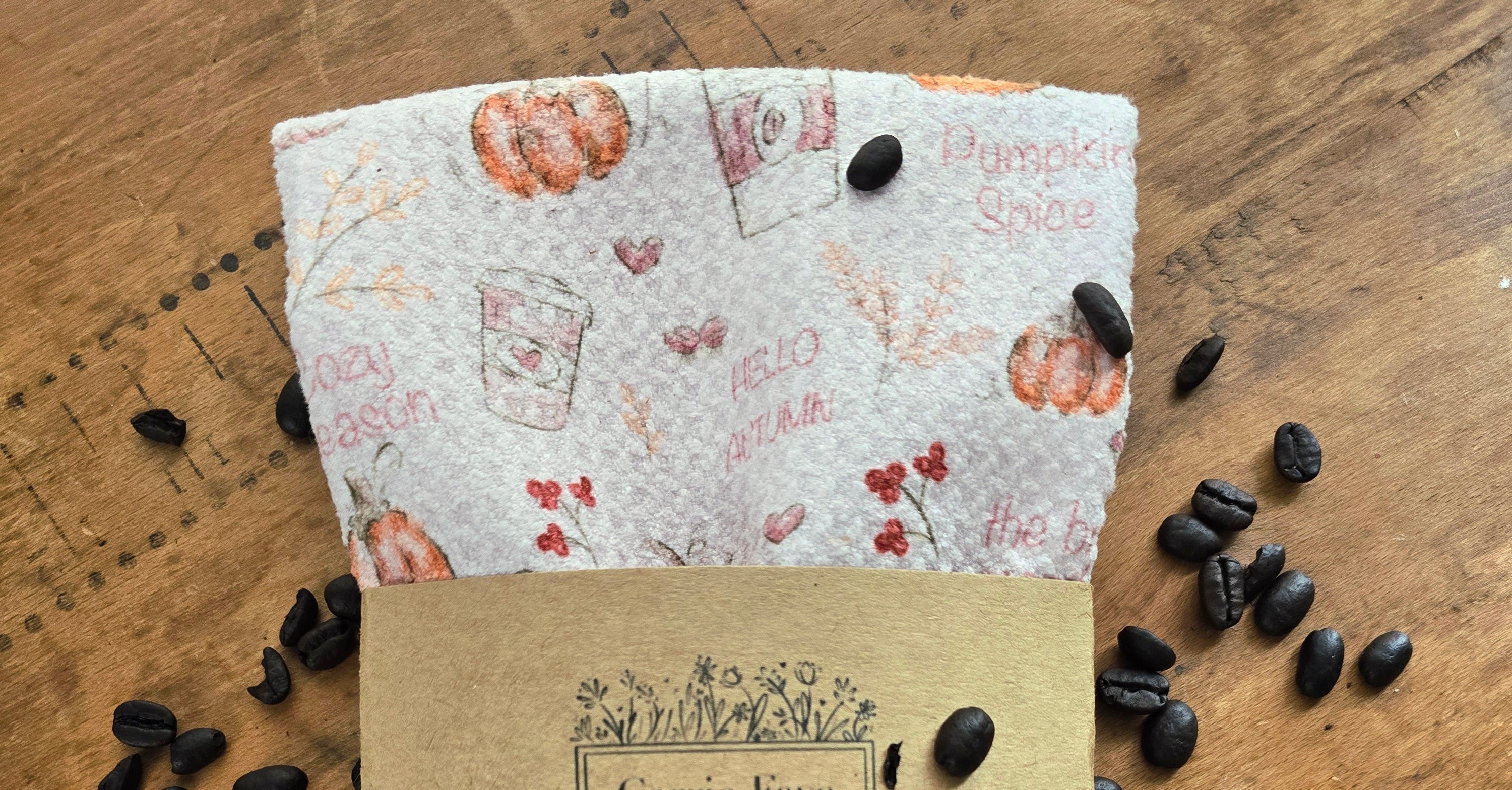 A folded microfiber towel with a fall-themed print of pumpkins, coffee cups, and text like 'Pumpkin Spice' and 'Hello Autumn,' wrapped in a kraft paper band labeled Carrie Faye, placed on a wooden surface with scattered coffee beans.