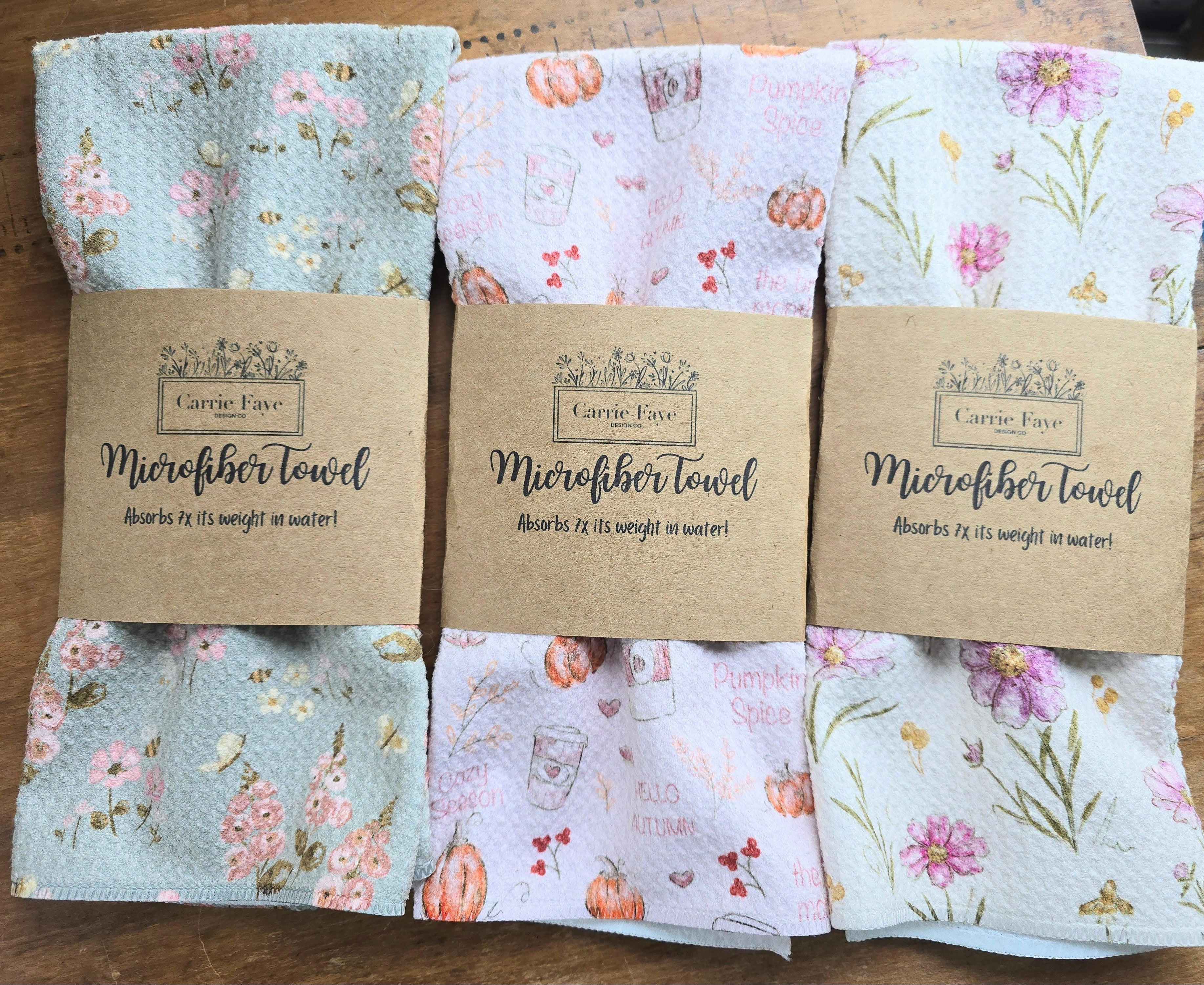three folded microfiber towels in different floral and botanical patterns, each wrapped with a brown kraft paper band labeled 'Carrie Faye Microfiber towel'.